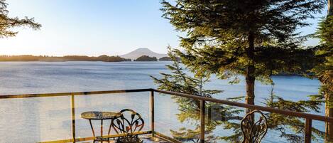 A deck with two metal chairs and a small table, overlooking a calm lake with a distant mountain and trees.