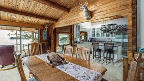 Un intérieur en bois comprenant une table à manger, des chaises, une tête de cerf accrochée au mur et un coin cuisine avec vue sur le lac.