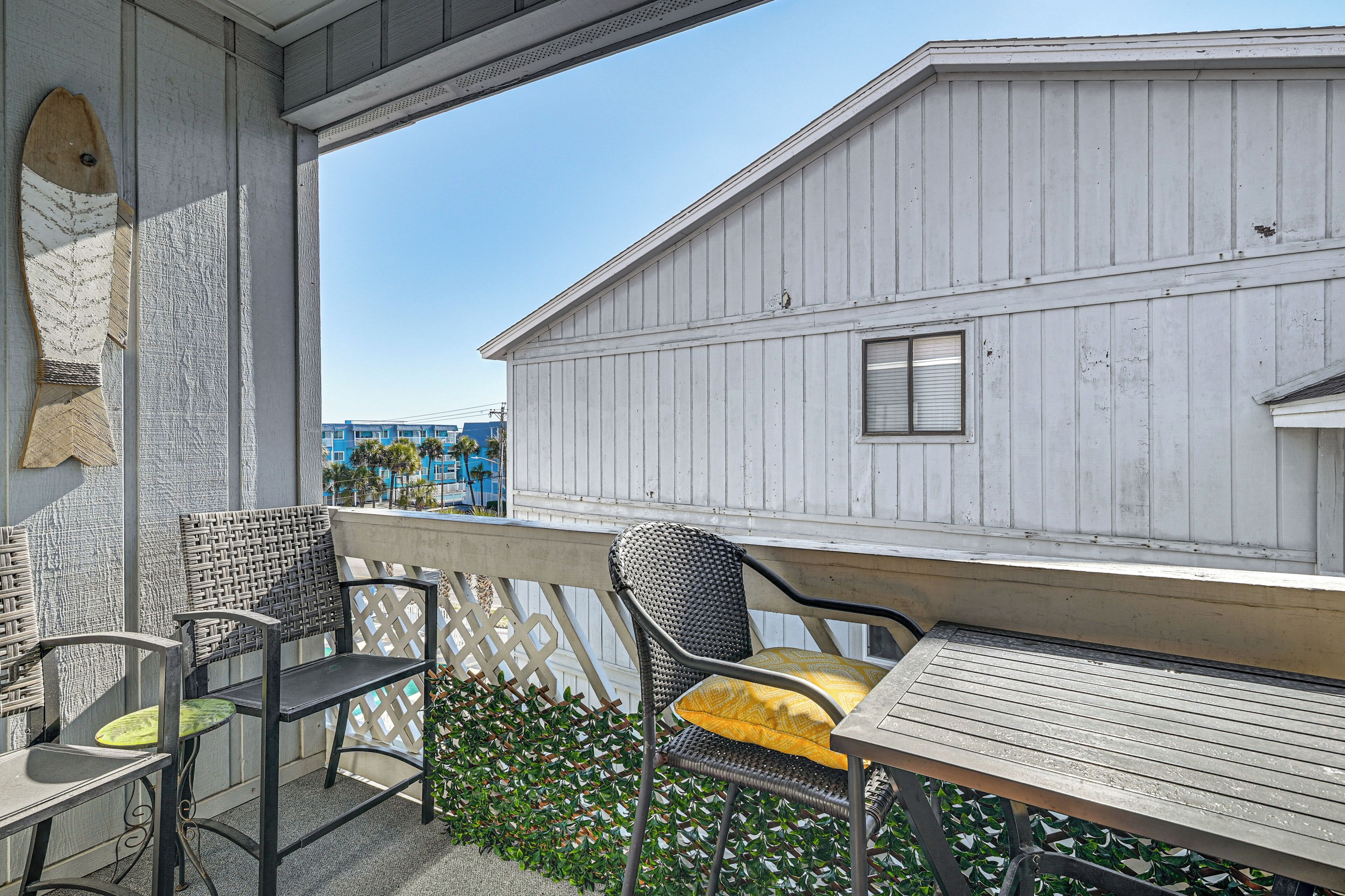 A balcony with wicker chairs and a wooden table, overlooking a building and a clear sky.