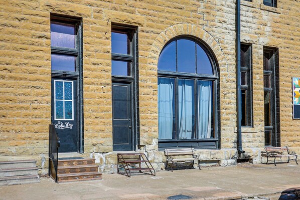 Un edificio histórico con amplias ventanas de arco, una puerta negra con un letrero y una banca en el exterior.