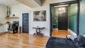 Un espacio habitable amplio con piso de madera, un sofá negro, una mesa de comedor con sillas, una cocina con horno y una puerta que da a otra habitación.
