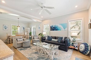 A spacious living room with a glass coffee table, a dark sofa, striped chairs, and a ceiling fan. - Freestanding 3br with tiki bar and pool (Pensacola)