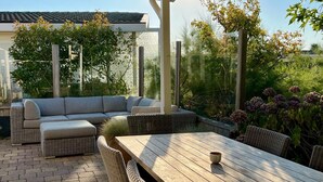 A covered patio with a wooden table, wicker furniture, and a view of surrounding greenery. - White Beach House (Breskens)