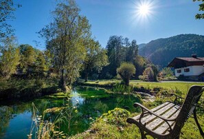 A serene lakeside setting with a wooden bench, a house in the background, and a clear sky.