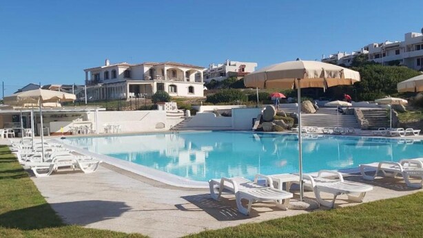 A swimming pool with lounge chairs, umbrellas, and a hillside with buildings. - Apartment With sea View and Great Roof Terrace (Santa Teresa di Gallura)