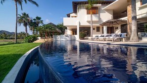 A modern resort with a large swimming pool, palm trees, and a two-story building with outdoor seating. - Casa Serena in Punta Mita (Punta de Mita)