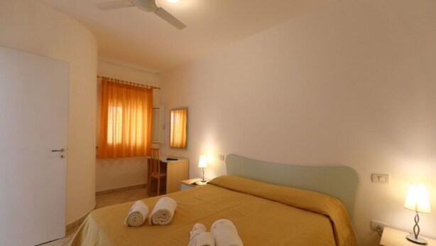 A hotel room with a bed, a desk, a chair, a nightstand with a lamp, and a ceiling fan. - Studio for Couples in Capo Vaticano With Pool (Ricadi)