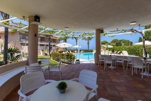 Ein überdachter Außenbereich mit weißen Stühlen und Tischen, einem Pool und Blick aufs Meer.