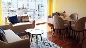 Un soggiorno con divano, poltrone, tavolino e zona pranzo con sedie e tavolo. Ampie finestre offrono vista sugli edifici esterni.