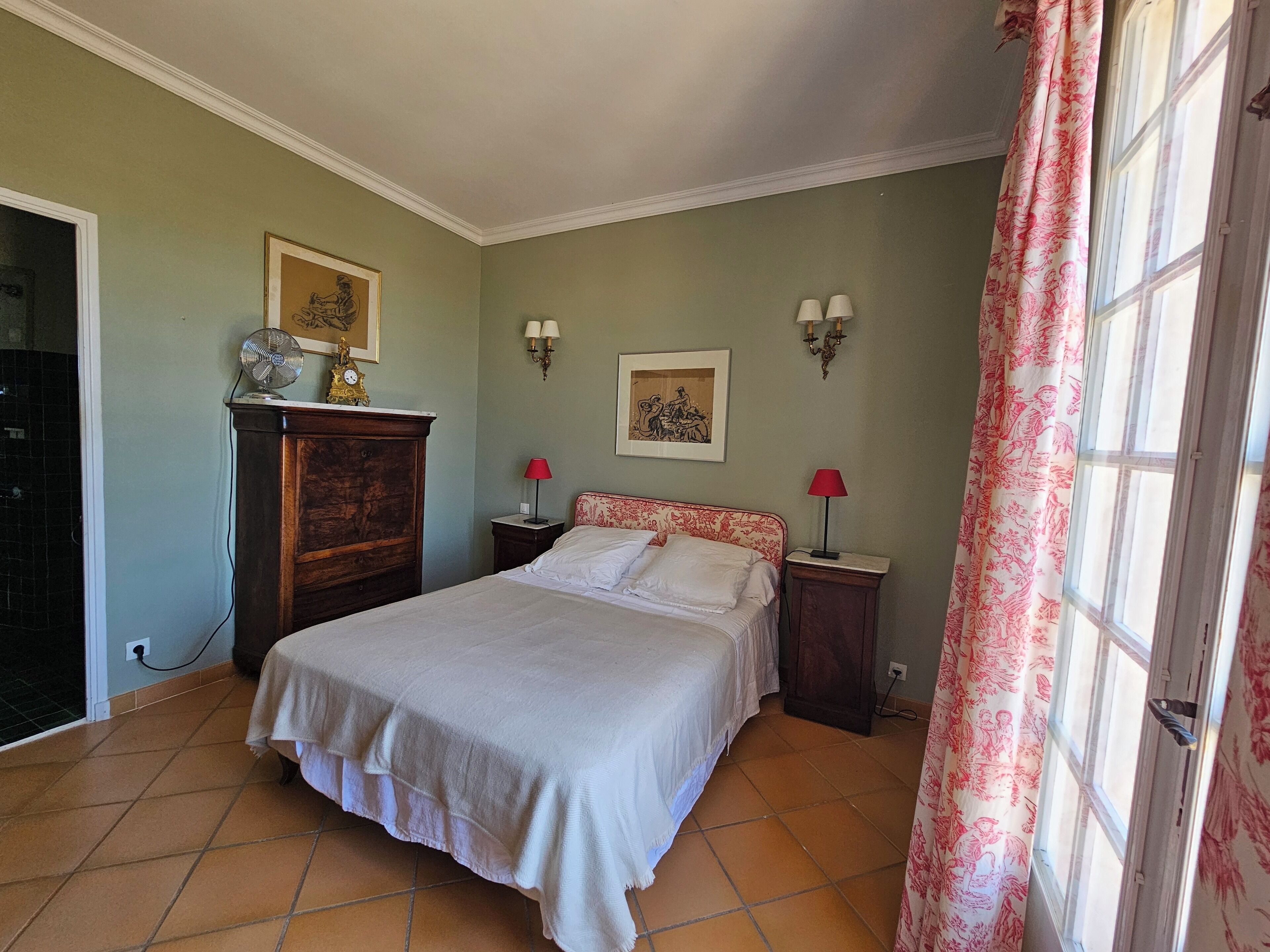Ein Schlafzimmer mit einem Bett, zwei Nachttischen, einer Kommode, einem Ventilator und einem Fenster mit Vorhängen.