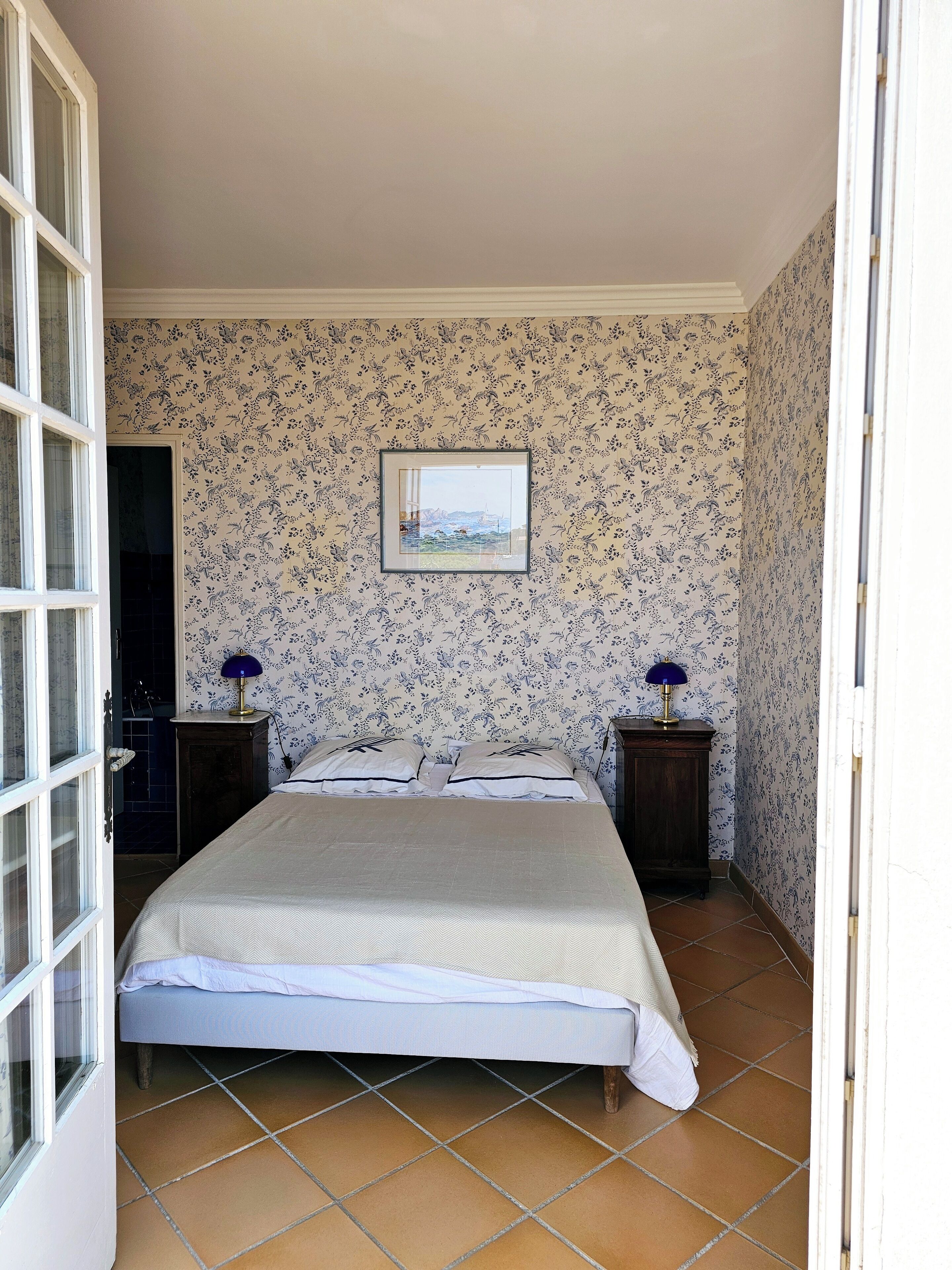 A bedroom with a bed, two bedside tables, a framed picture on the wall, and a door leading to another room.