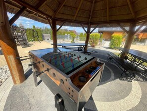An outdoor foosball table under a wooden shelter with a thatched roof, surrounded by picnic tables and greenery. - Holiday Home With a Terrace, Close to the Lake (Lubrza)