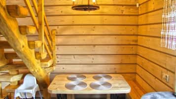 Interior de una cabaña de madera con una mesa de comedor, sillas, una lámpara colgante y una escalera de madera.