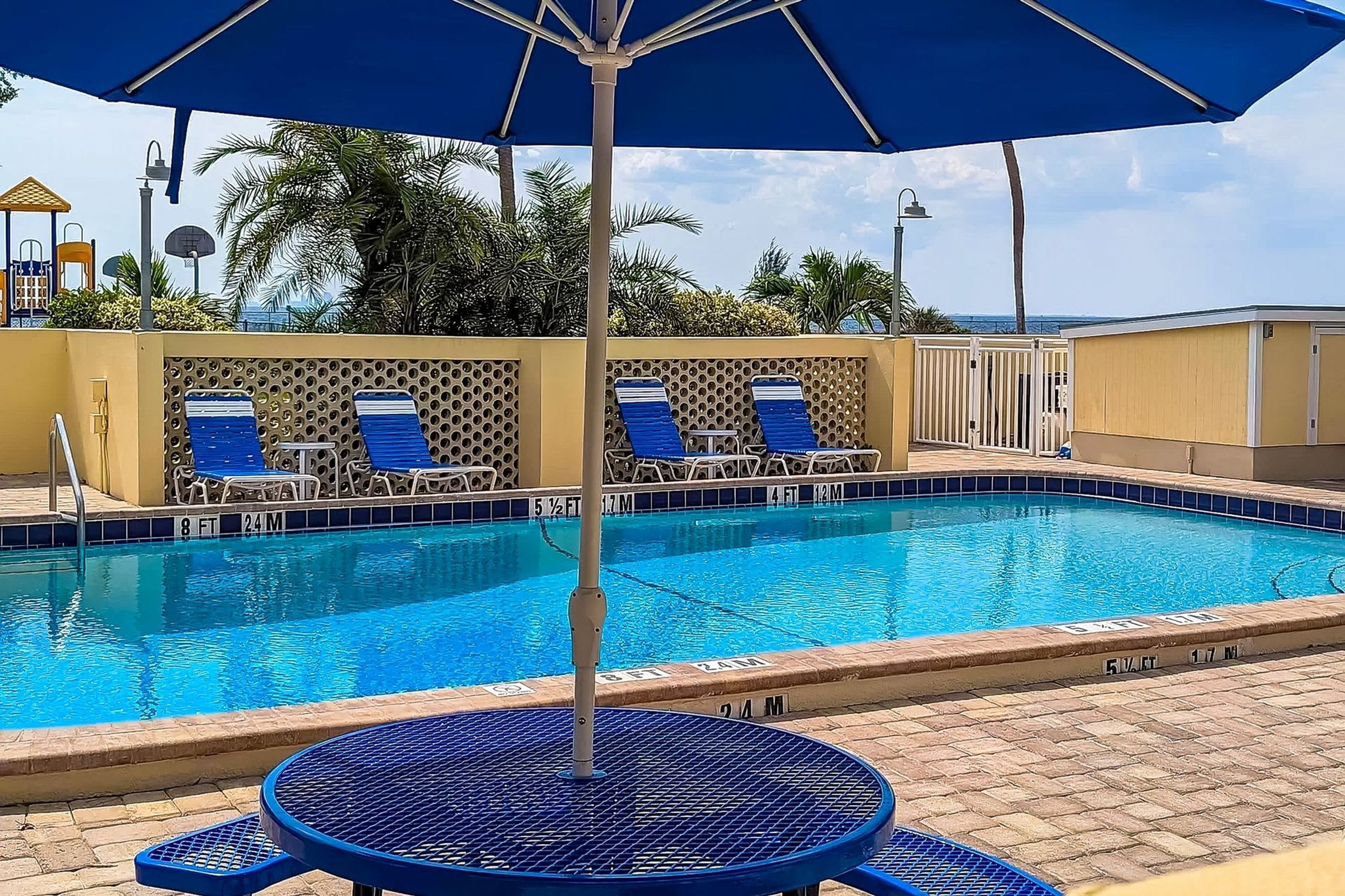 A swimming pool with blue chairs and an umbrella, surrounded by palm trees and a view of the ocean.