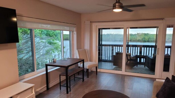A living room with a TV mounted on the wall, a dining table with chairs, a sofa, and a large window with a view of a lake. - Waterfront Balcony Condo Beach Access Hot Tub (Ruskin)