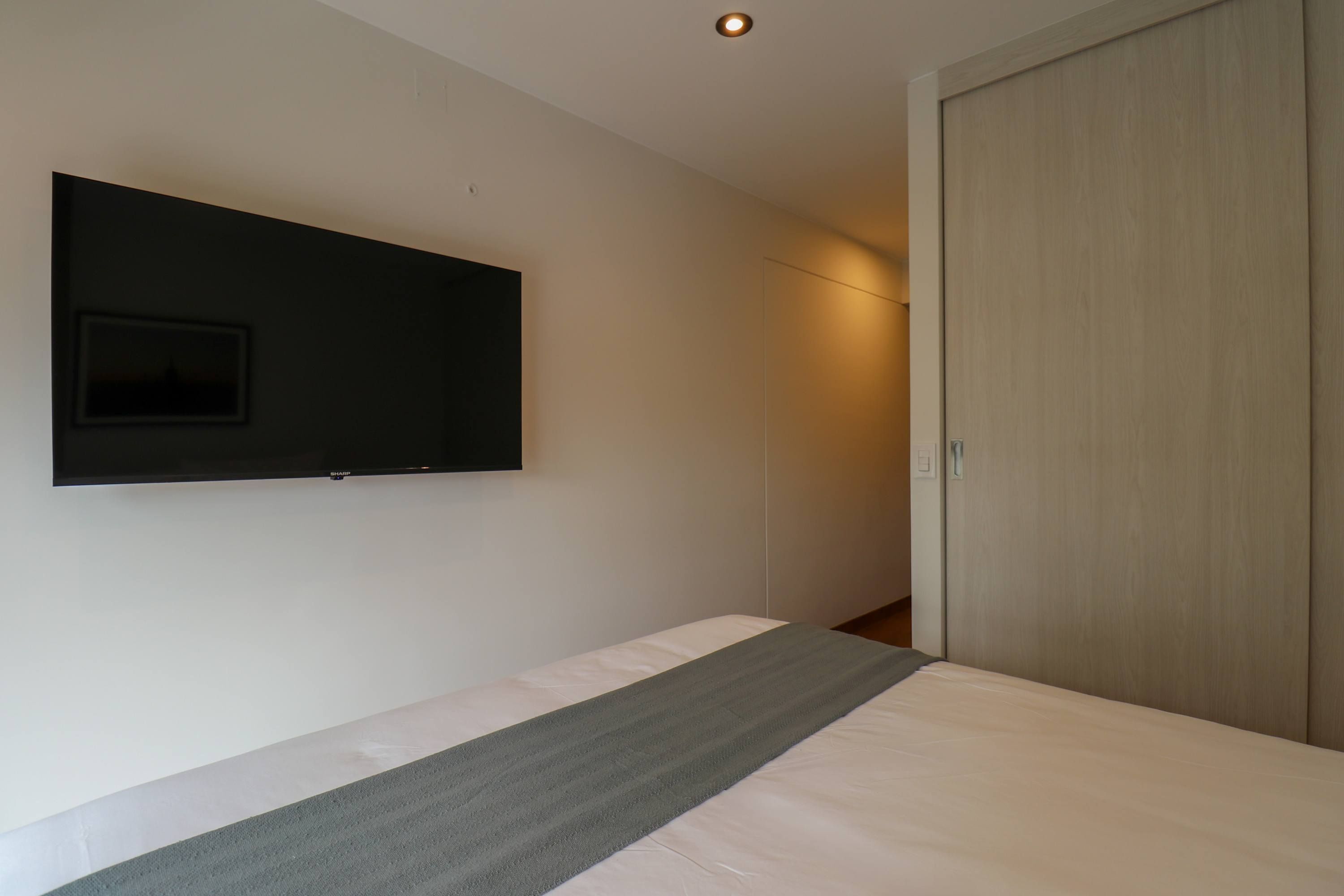 A bedroom with a bed, a flat-screen TV mounted on the wall, and a wooden wardrobe.