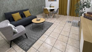 Un salon comprenant un canapé, une table basse et un coin repas avec des chaises. On y trouve également une plante et un tableau d’affichage au mur.