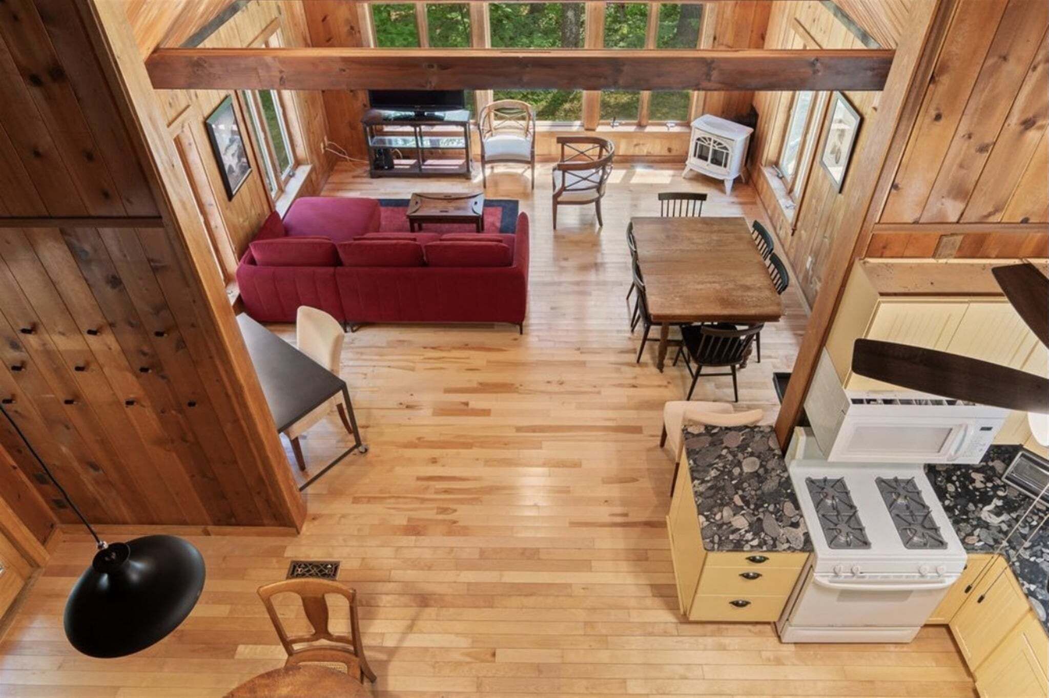 A wooden cabin interior with a dining area, a sofa, a kitchen with a stove, and a large window overlooking greenery.