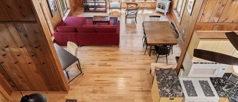 A wooden cabin interior with a dining area, a sofa, a kitchen with a stove, and a large window overlooking greenery.