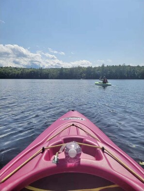 A pink kayak with a bottle and a whistle on the bow, heading towards another kayak on calm water with a forested shoreline in the background.