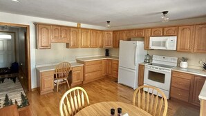 A compact kitchen with wooden cabinets, a refrigerator, a microwave, and a stove. There is a dining table with chairs and a small rug. - Upta Camp Limit 8 3 Bedroom Cabin (East Baldwin)