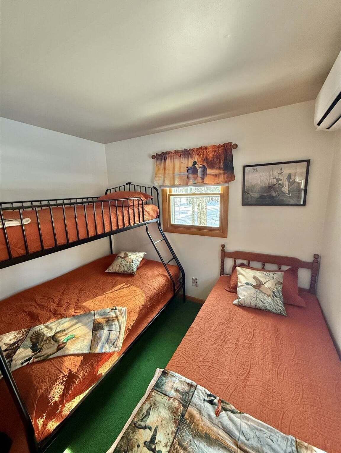 A small, compact room with two bunk beds, a window with a view of the outdoors, a framed picture on the wall, and a small air conditioning unit.
