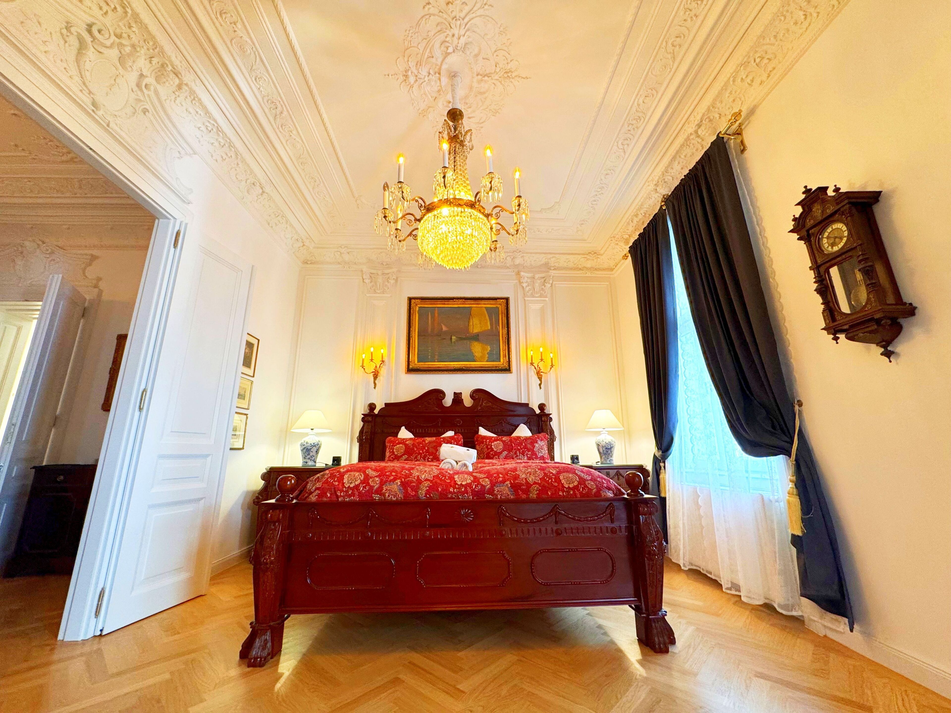 A spacious bedroom with a large wooden bed, ornate ceiling, and a classic chandelier.