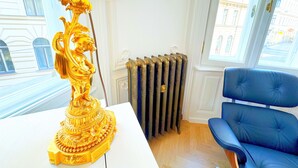 Quarto com abajur dourado, radiador, cadeira azul e janela com vista para edifícios.