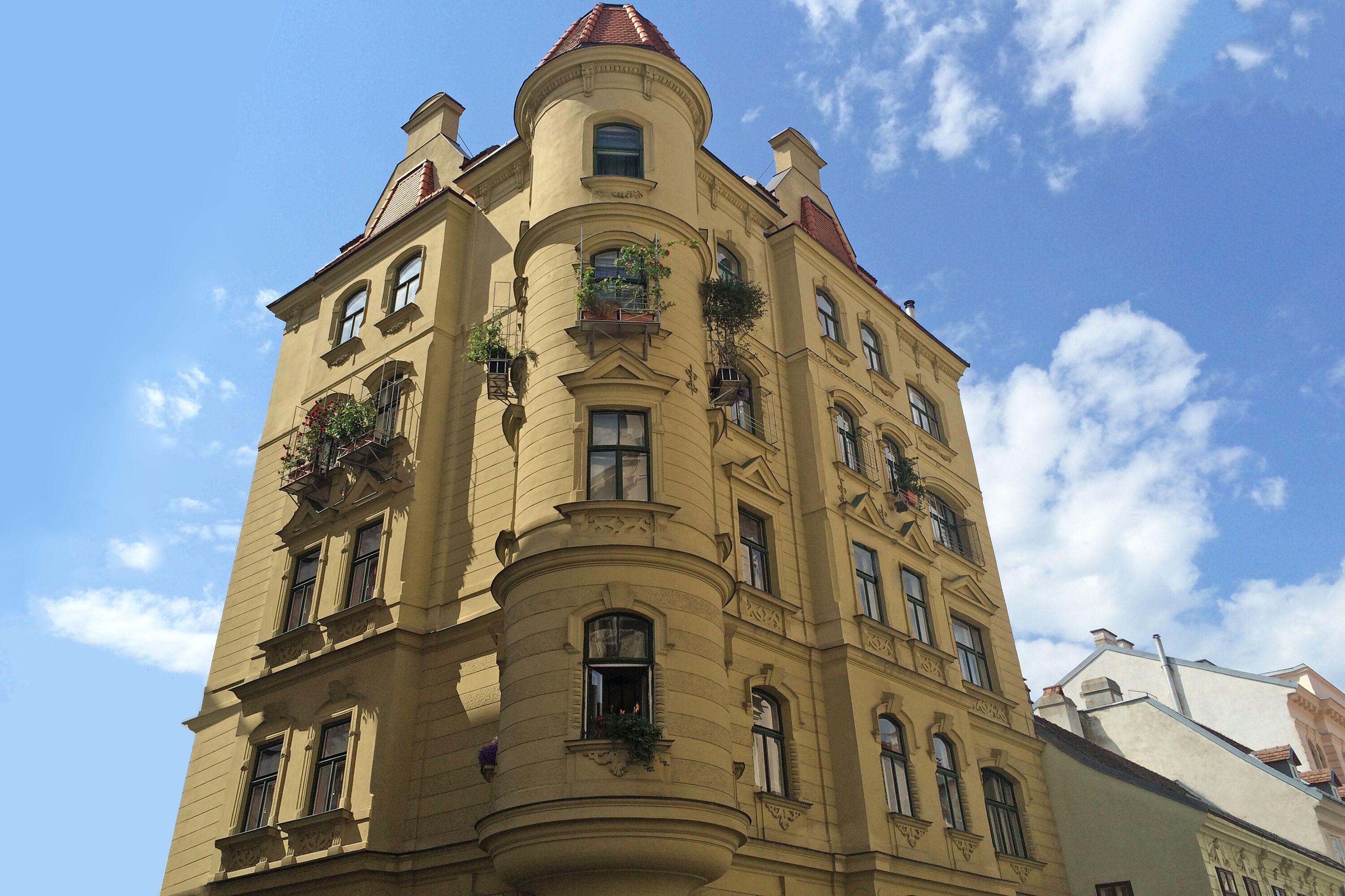 Ein mehrstöckiges Gebäude mit abgerundeter Ecke, zahlreichen Fenstern und Balkonen mit Pflanzen.