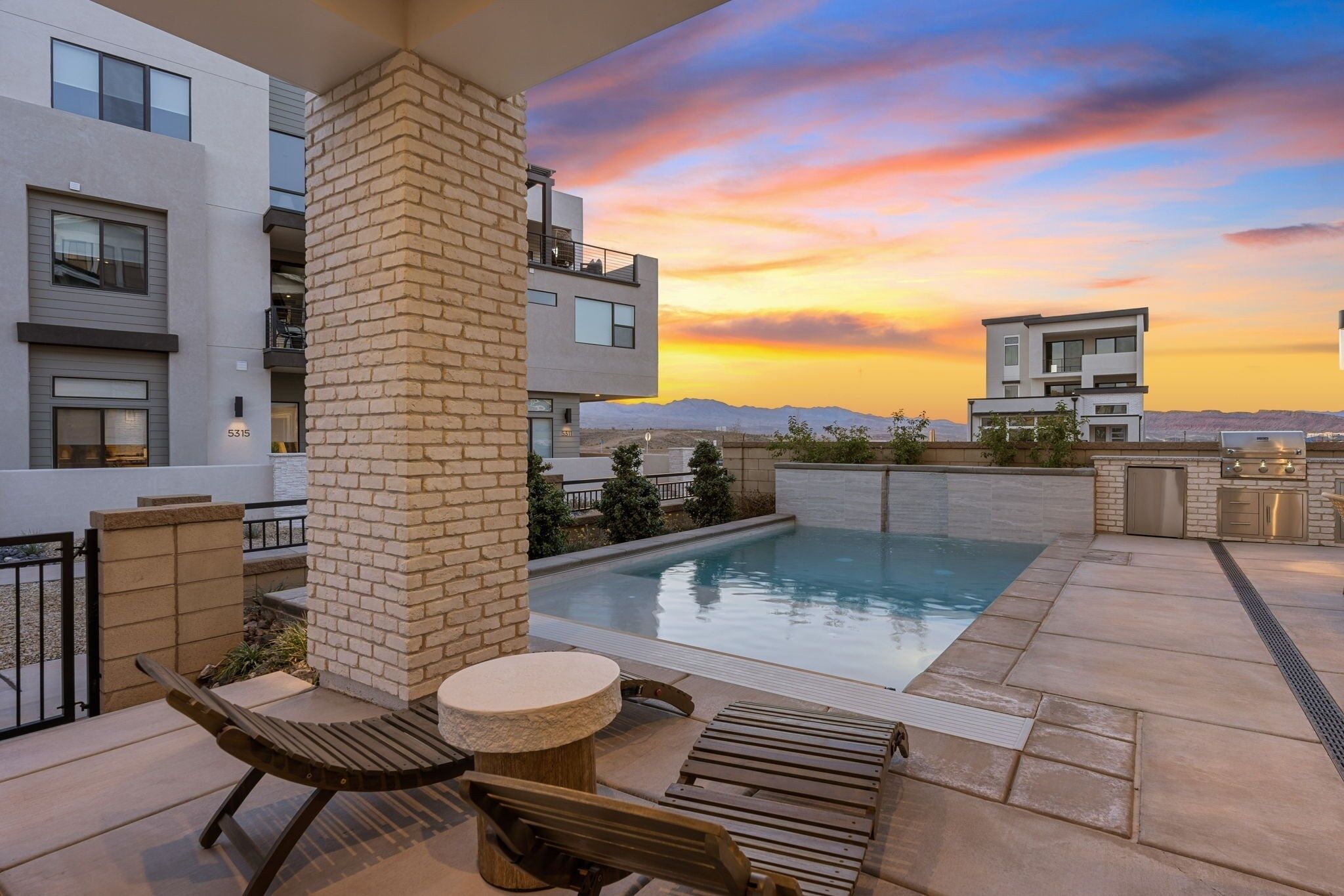 A rooftop pool area with a stone pillar, lounge chairs, and a view of the mountains at sunset.