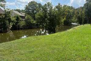 A river with a grassy bank, trees, and a building in the background. - Skinny Dippin 2 Bedroom Cabin (Pigeon Forge)