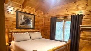 A wooden cabin bedroom with a bed, a framed picture on the wall, and a window with curtains. - Skinny Dippin 2 Bedroom Cabin (Pigeon Forge)