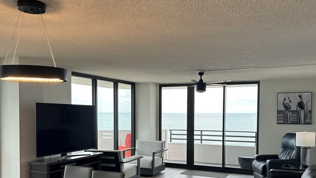 Een moderne woonkamer met een glazen tafel, stoelen, een bank, een televisie en uitzicht op zee door grote ramen.