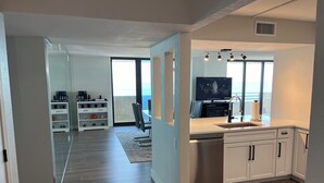 Een moderne keuken met een vaatwasser, een spoelbak en een televisie. Er is een eethoek met een tafel en stoelen, en een keukeneiland met een koffiezetapparaat. De kamer heeft grote ramen en uitzicht op de oceaan.