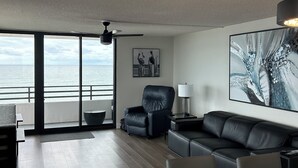 Een moderne woonkamer met een groot raam, een zwarte leren bank, een glazen salontafel en een plafondventilator.