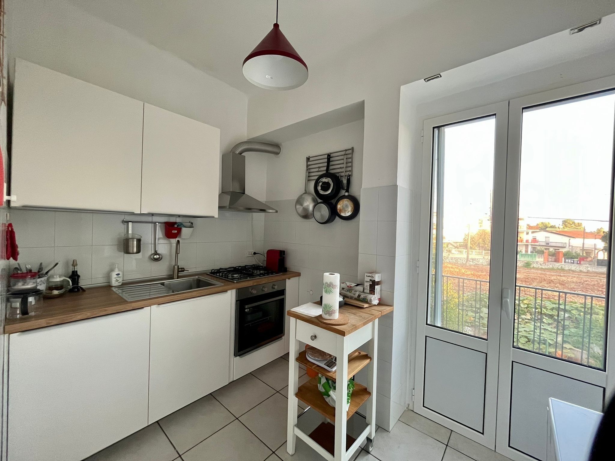 A compact kitchen with white cabinets, a gas stove, and a wooden countertop. There is a window with a view of a garden and buildings outside.