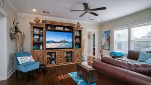 Una sala de estar con un televisor grande, un mueble de entretenimiento de madera, un sillón azul, un sofá marrón y un ventilador de techo.