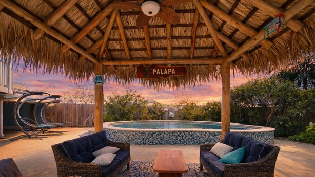 Un área de estar al aire libre techada con mobiliario de mimbre, piscina y un letrero que dice "PALAPA".