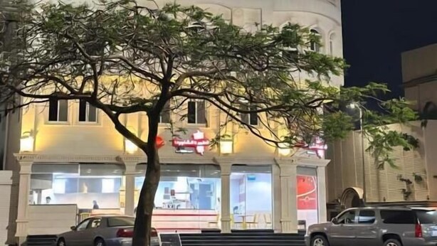 Un edificio con un letrero en árabe e inglés, un árbol en la parte delantera y automóviles estacionados afuera.
