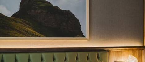 Une chambre d’hôtel avec un lit, un tableau représentant une montagne accroché au mur, une tête de lit recouverte d’un tissu vert et une table de chevet avec une lampe.
