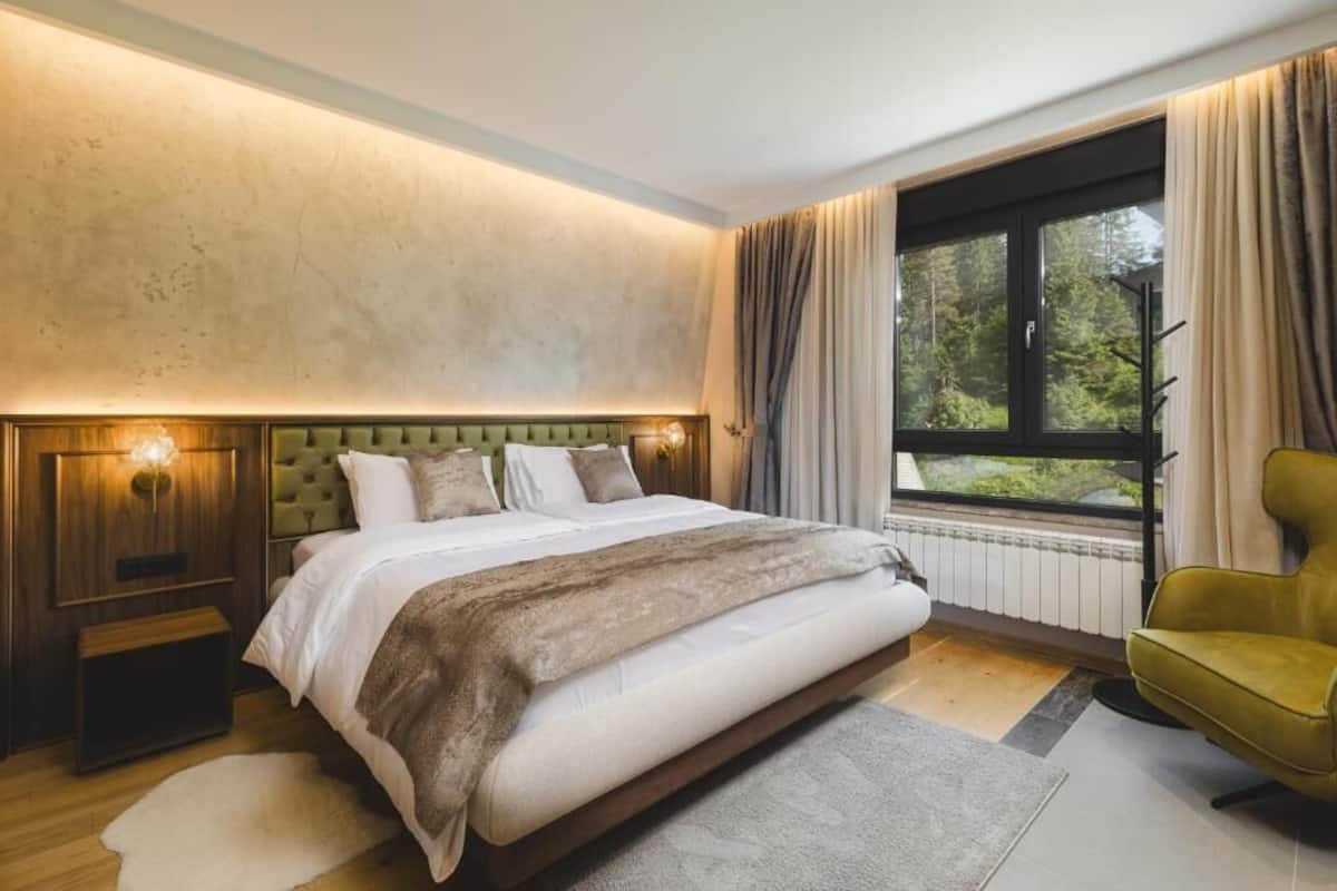 A bedroom with a large bed, a nightstand, a chair, a window with curtains, and a view of trees.