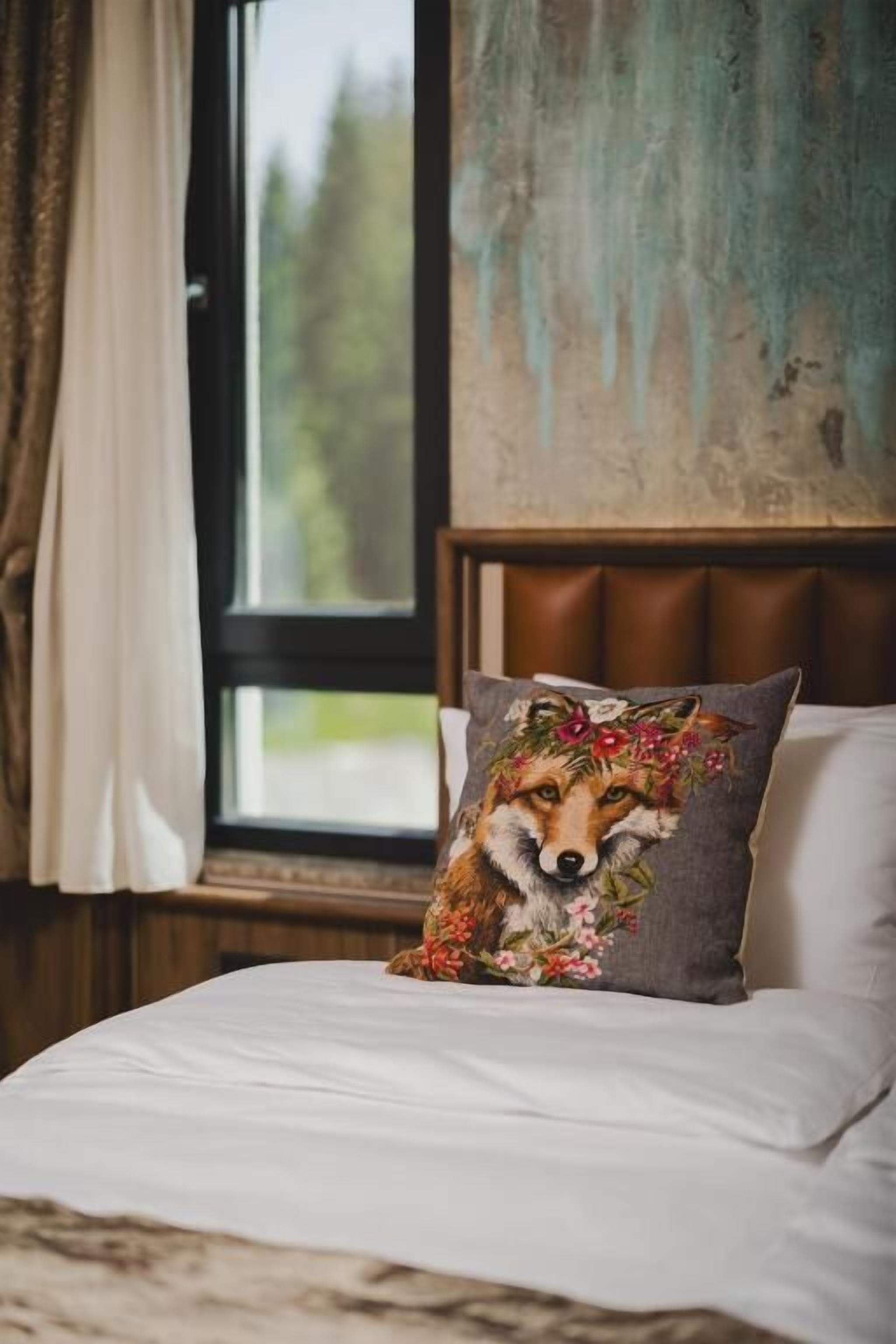 Una cama con un almohadón de diseño de zorro, una ventana con cortinas y vistas al verdor exterior.