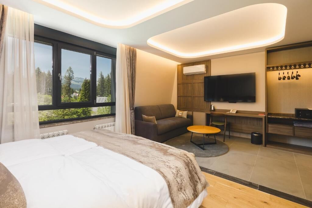A modern hotel room with a bed, a sofa, a small table, a TV, and a view of the outdoors.