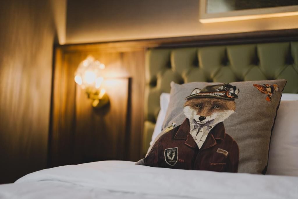 Una cama con un cojín que muestra un zorro con uniforme, un cabecero capitonado y una lámpara de pared.