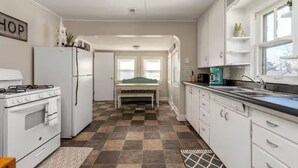 Cocina compacta con armarios blancos, estufa y refrigerador. Hay un banco en el centro de la habitación y una ventana a la derecha.