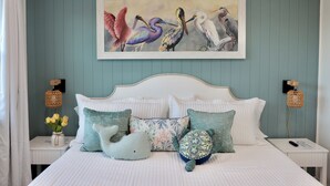 Quarto com cama, almofadas, pintura de pássaros na parede e duas mesas de cabeceira com abajures.