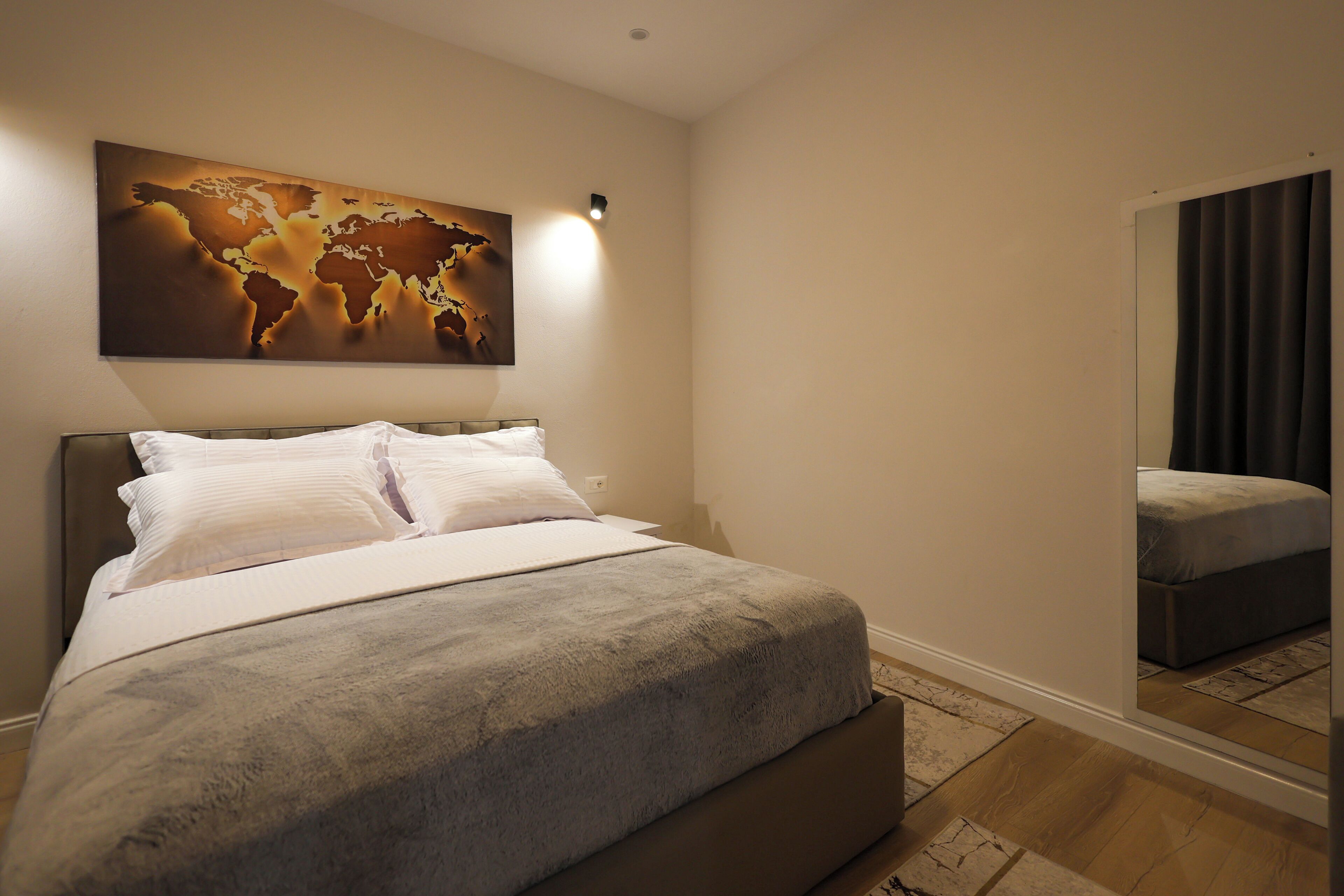 A neatly made bed with white linens, a world map artwork above it, and a large mirror reflecting the room.