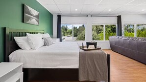Een moderne slaapkamer met een groot bed, een grijze bank en uitzicht op groen door grote ramen.