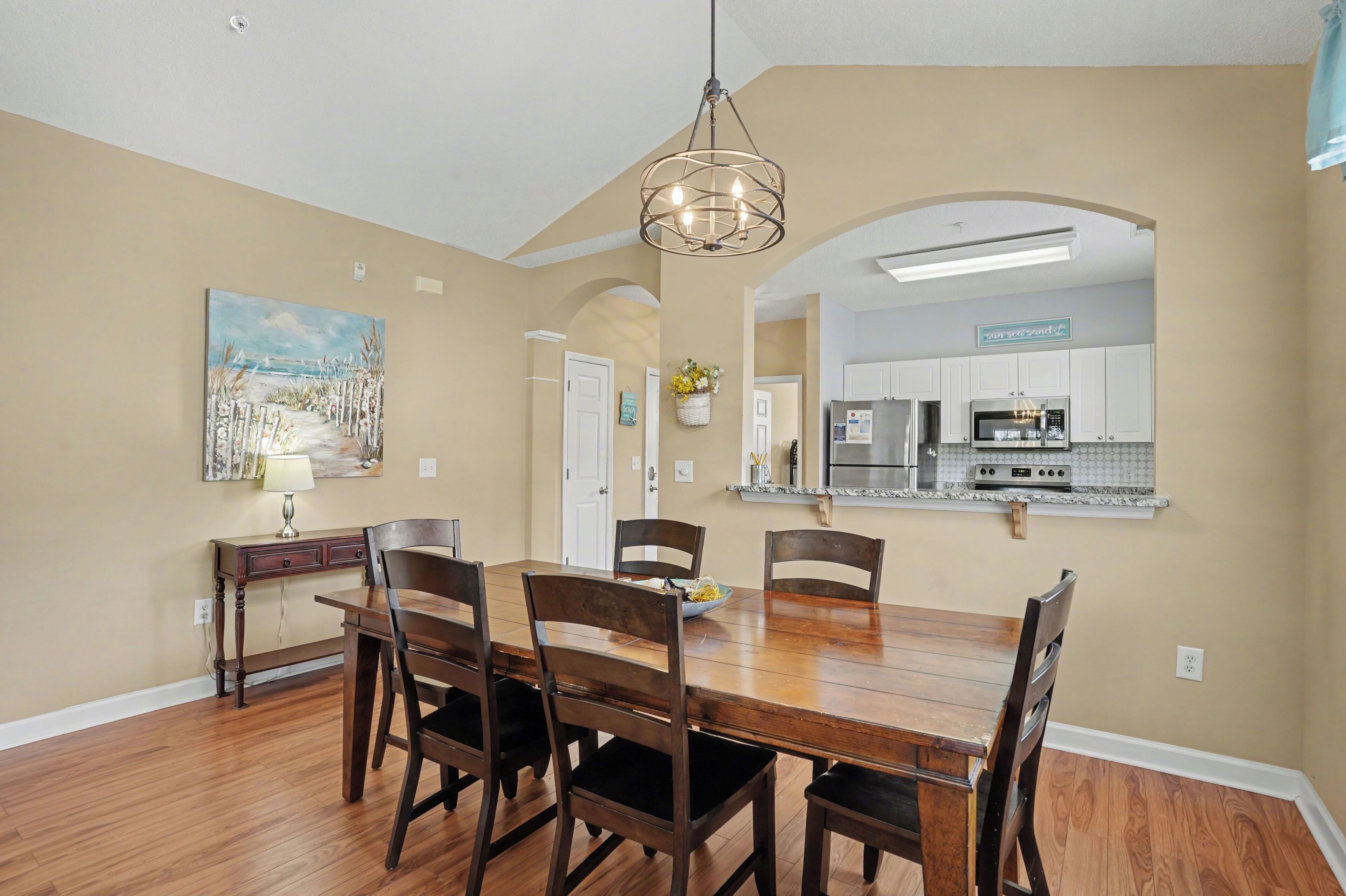 A dining room with a wooden table and chairs, a chandelier, and a kitchen in the background.