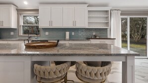 Uma cozinha moderna com armários brancos, bancada de granito e uma ilha grande com duas cadeiras estofadas.
